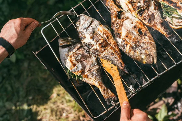 Ruce muže s vidličkou a dřevěnou špachtlí v popředí. Vaří lahodné a čerstvé grilované ryby s citrónem na grilu na zahradě v létě. Výběr snímku makra zaměření — Stock fotografie