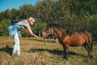 Uzun saçlı, pembe tişörtlü, açık mavi kotlu, kot ceketli, uzun saçlı, güzel, sarışın bir kadın eğilmiş ve elmanın arka planındaki kahverengi midilliye elma yediriyor.