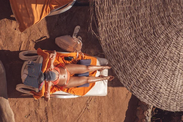 Schöne junge Mädchen in einem gestreiften orangefarbenen Badeanzug liegt auf einer Sonnenliege unter einem Strohschirm und sonnt sich — Stockfoto