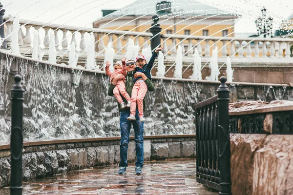 Moscú, Rusia - 7 de julio de 2017. Un padre feliz con gafas de sol sostiene a dos hijas en sus brazos. se ríen y se regocijan. En el fondo de la fuente de la ciudad — Foto de Stock