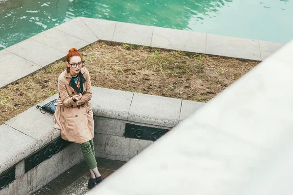 Moscou, Rússia - 7 de julho de 2017. Uma jovem hipster pensante com uma capa de chuva bege e calças curtas verdes e óculos fica em um passeio em um parque perto da praça vermelha e do Kremlin. Ela fuma um cigarro. — Fotografia de Stock