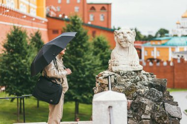 Moskova, Rusya - 7 Temmuz 2017. Omzunda siyah bir çanta ve elinde şemsiye olan bej ceketli ve pantolonlu bir adam, durup aslan şeklinde heykele bakar. Şeyde...