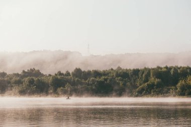 Balıkçı teknede balık tutuyor. Sabahın erken saatlerinde, ön planda nehir ve arka planda çalılar ve ağaçlar olan yeşil bir banka olan ilkbahar manzarası. Uzak plan yoğun bir sabah sisinde boğuldu.