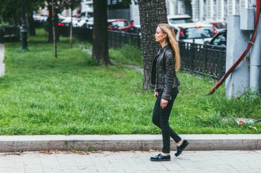 Moskova, Rusya - 7 Temmuz 2017: Deri ceketli, uzun saçlı, beyaz bir kadın, ağaçların ve yeşil çimlerin arkasında parkta yürüyor.