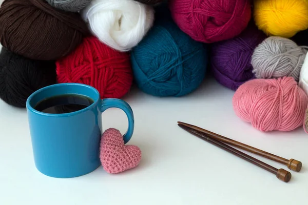 Cup of coffee and knitting needles with colour balls of yarn on white table. with pink heart of wool yarn