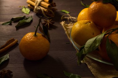 Öndeki yeşil dalda lezzetli bir mandalina. Masanın üzerinde olgun meyveler var. Taze turuncu mandalinalar ve koyu ahşap zemin üzerinde cam bir tabakta yeşil yapraklar. alçak tuş.