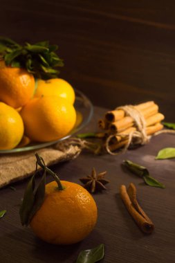 Dikkat çekmeden. Ahşap arka planda yeşil yapraklı taze turuncu mandalinalar. Olgun meyveler karanlık masada tarçın ve baharatlarla.