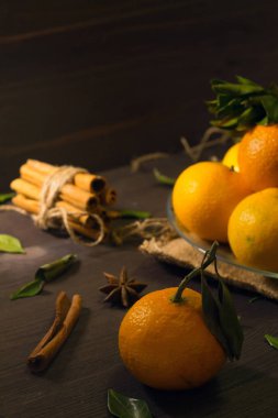 Masanın üzerinde olgun meyveler var. Öndeki yeşil dalda lezzetli bir mandalina. Taze turuncu mandalinalar ve cam bir plaka içinde yeşil yapraklar. Koyu ahşap arka planda baharatlı. Dikkat çekmeden. dikey