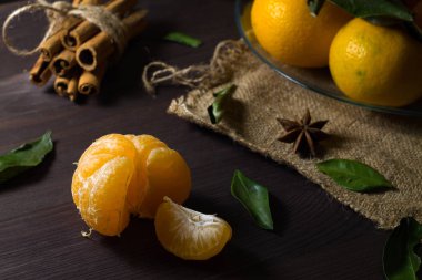 Dikkat çekmeden. Ahşap masada soyulmuş mandalina. Ahşap arka planda yeşil yapraklı taze turuncu mandalinalar. Olgun meyveler karanlık masada tarçın ve baharatlarla.