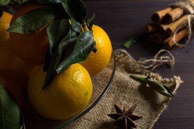 Taze ve olgun portakal mandalinaları ve mandalinalar cam tabakta yeşil yapraklar, tarçın çubukları ve ahşap arka planda baharatlar. alçak tuş.