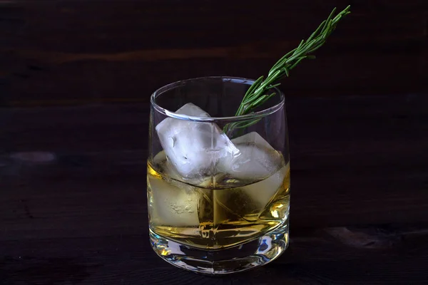 drink in a glass with ice and a sprig of rosemary on a dark wooden background. glass of whiskey or cocktail with ice. glass of lemonade with pieces of ice