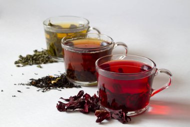 Beyaz arka planda siyah, kırmızı ve yeşil yapraklı üç bardak çay. Kuru çiçekli kırmızı Hibiscus çayı. Sağlıklı bir yaşam tarzı. Yeşil sıcak çay. Siyah sıcak çay. Sıcak içecekler.