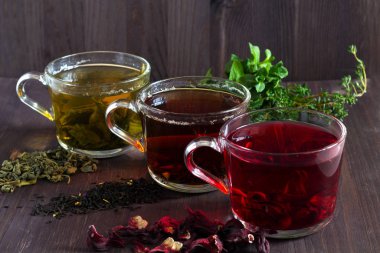 Üç bardak siyah, kırmızı amber çiçeği ve kuru yapraklı yeşil bitkisel çay ve koyu ahşap zemin üzerinde taze nane yaprakları. Sağlıklı bir yaşam tarzı. Sıcak içecekler. yatay yönelim.