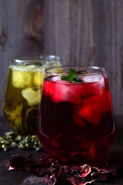 Ahşap bir masada bir bardak buzlu kırmızı amber çayı ve kuru çiçekler. Sağlıklı bir yaşam tarzı. Koyu ahşap zemin üzerinde nane yaprakları olan bir bardak buzlu yeşil bitki çayı. Soğuk içecekler. düşey yönelim.