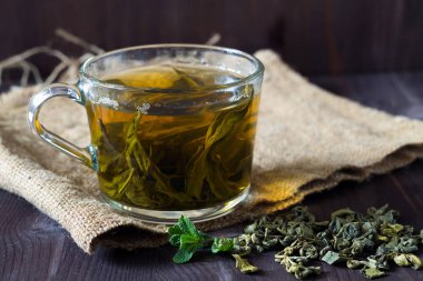 Bir bardak yeşil bitkisel çay, kuru yapraklar, yeşil nane yaprakları ve koyu ahşap bir arka planda çuval bezi. Sıcak içecekler. Yatay oryantasyon. kopyalama alanı. Sağlıklı yaşam tarzı.