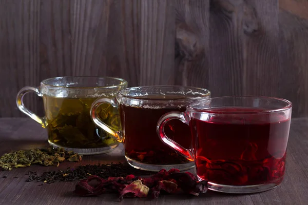 Healthy lifestyle.Hot drinks.Three glass cups with black, red and green tea with dry leaves and flowers on a dark wooden background. Red hot Hibiscus tea. Black hot tea. Green hot tea on wooden table.