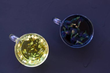 Siyah masa manzaralı iki fincan kuru çiçekli sıcak çay. Sıcak kelebek çayı ve yasemin çayı. Sağlıklı bir yaşam tarzı. Siyah arka planda mavi ve sarı renkli cam fincanlar. Sıcak içecekler.