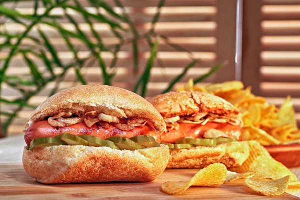 Perro caliente a la parrilla o salchicha con cebolla frita y pepinos marinados. Patatas fritas. Última comida rápida clásica. Luz cálida sobre mesa de madera . — Foto de Stock