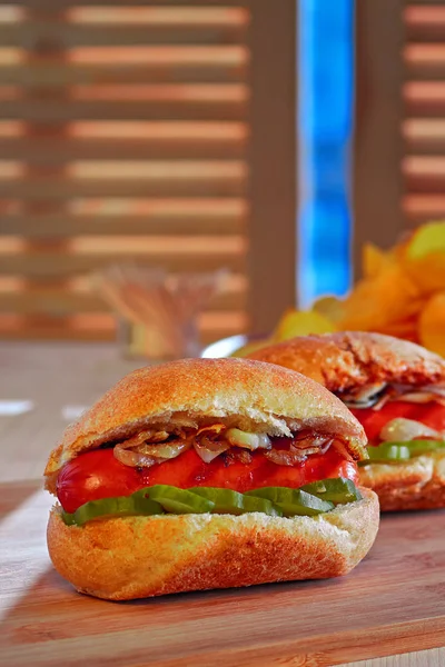 Perro caliente a la parrilla con cebolla frita y pepinos marinados. Patatas fritas. Comida rápida en mesa de madera. Luz del día brillante . — Foto de Stock