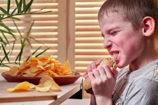 Băiatul cu poftă de mâncare mănâncă un hot dog delicios. Copilul flămând își rupe dinții de bună voie și mestecă hotdog. Fast food . — Fotografie, imagine de stoc