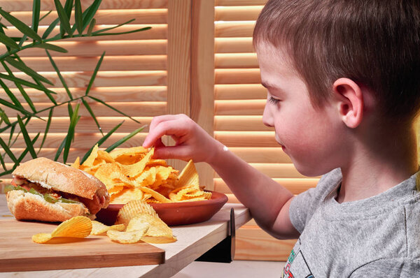 Мальчик ест вкусные чипсы в кафе. Ребенок держит чипсы в руке. На столе лежит укушенный хот-дог. Фаст-фуд
.