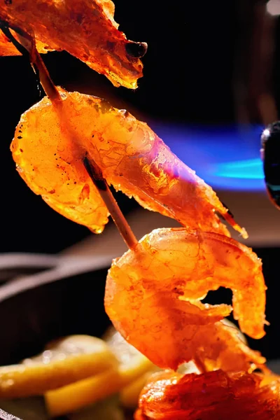 Shrimps on wooden skewer fry with cooking gas torch flomber. Preparation delicious fresh sea food with professional chef equipment on background frying pan with garnish. Vertical photo. — Stock Photo, Image