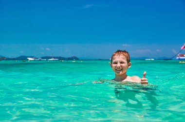 Çocuk (10 yaşında) Turkuaz su tropikal Denizde yüzme. Başparmak gösterilen süre gülen çocuk. Parlak mavi gökyüzü ve parıldayan deniz deniz manzarası. Kopya alanı.