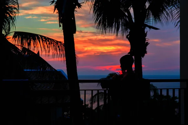 Criança silhueta ao pôr-do-sol. Menino fica na varanda em meio ao pôr-do-sol do mar vermelho e palma. Pôr-do-sol dramático sobre o mar tropical . — Fotografia de Stock