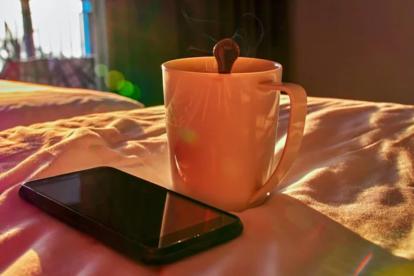 Concept confortable. Tasse blanche et smartphone noir sur le lit dans la lumière du soir ensoleillée de la fenêtre ouverte. L'été sous les tropiques . — Photo