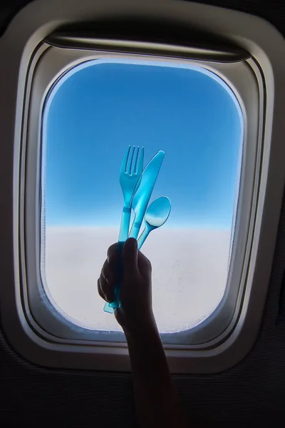 Концепція харчування у пасажирському літаку під час польоту. Дитяча рука тримає пластикову ложку, виделку, ніж проти вікна з хмарами і блакитним небом . — стокове фото