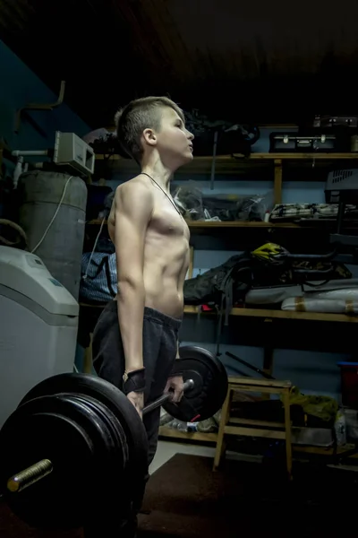 Pojke (12 år) lyfter en skivstång i garaget. Styrketräning hemma. Påfrestade muskler, dödlyft — Stockfoto