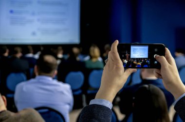 İş adamlarının elleri akıllı telefon tutarken ve resmi konferansta iş sunumuyla ekranın fotoğraflarını çekerken, kalabalık bir odada buluşurken..