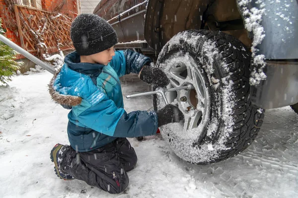 Child (10 years old) repairs a car. Boy screws a nut on car wheel, snowy winter day. Replacement wheels in the snow — Stok fotoğraf