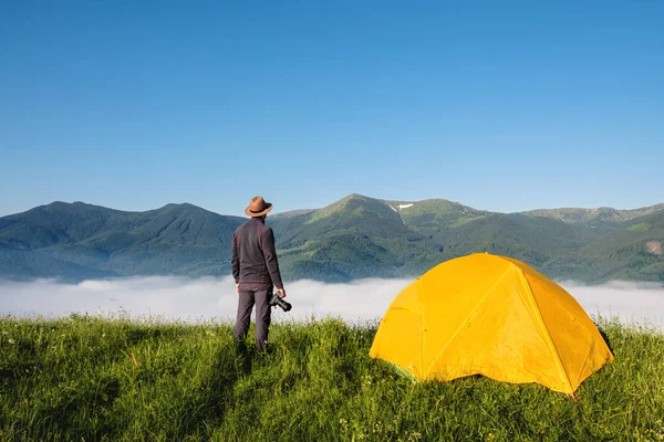 Man photographer with photo camera shooting foggy mountains summer landscape near camping tent. Travel and Lifestyle concept. — Stock Photo, Image