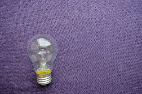 A round, ordinary, non-economical incandescent bulb with a transparent socle background of purple cloth.