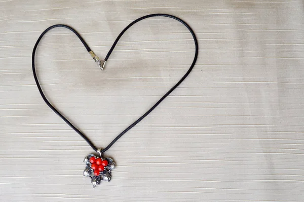 Women\'s necklace with a silver pendant with red circles in the form of a heart to the day of St. Valentine made of black thread on a beige background.