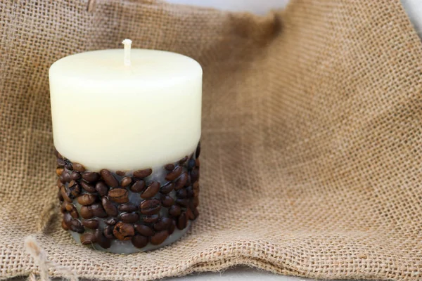 Wax beautiful light beige candle with unflavored wick from below decorated with coffee beans on the background of old brown canvas fabric.