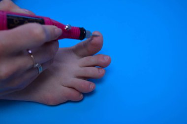 Mavi arka planda özel bir makinenin yardımıyla tırnağın tırnak işleme işleminin ayak parmaklarına pedikür yapma süreci