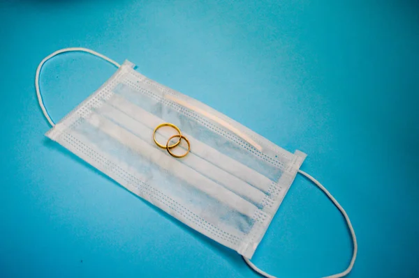 As alianças de casamento de ouro estão em uma máscara médica protetora descartável para proteção contra doenças mortais perigosas de micróbios e vírus pelo coronavírus covid-19 em um fundo azul — Fotografia de Stock