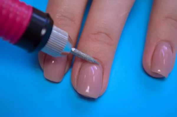 El proceso de hacer una hermosa manicura en los dedos de un dedo procesando una uña con una máquina especial en un salón de belleza de uñas sobre un fondo azul —  Fotos de Stock