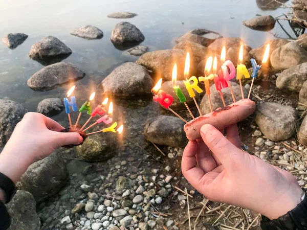 Burning happy birthday inscription made of holiday candles in the hands of a man and a woman opposite the water of the ocean lake river. Concept: birthday celebration in nature, outdoors — Stock Photo, Image