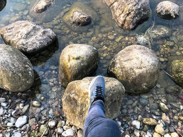 A foot in a sneaker steps on large round beautiful natural stones cobblestones in water, sea, lake, river. Concept: tourism, outdoor activities