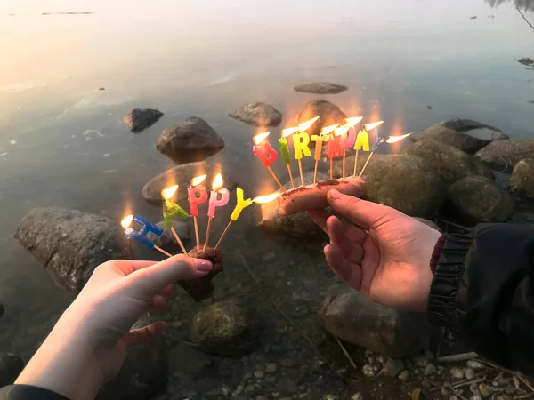 Pálení šťastný narozeninový nápis z svátečních svíček v rukou muže a ženy naproti vodě oceánu jezera řeky. Koncept: narozeninová oslava v přírodě, venku — Stock fotografie