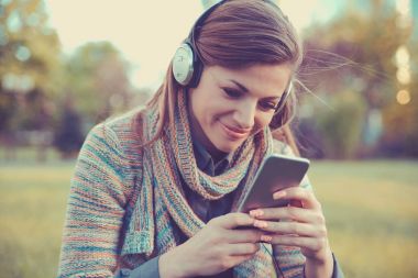 Mutlu kadın bir akıllı telefon videoları arka planda bir odaklanmamış binaları ile bir parkta oturan kulaklıklar ile izliyor