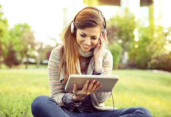Uśmiechnięta kobieta, słuchanie muzyki z tabletu i słuchawki siedzi na świeżym powietrzu w parku miejskim — Zdjęcie stockowe
