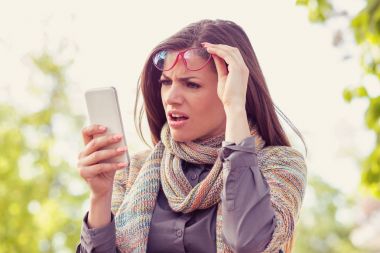 Annoyed upset woman in glasses looking at her smart phone with frustration while walking on a street on an autumn day   clipart