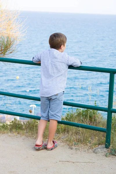 Menino fica perto de cerca de metal e olha para o mar — Fotografia de Stock