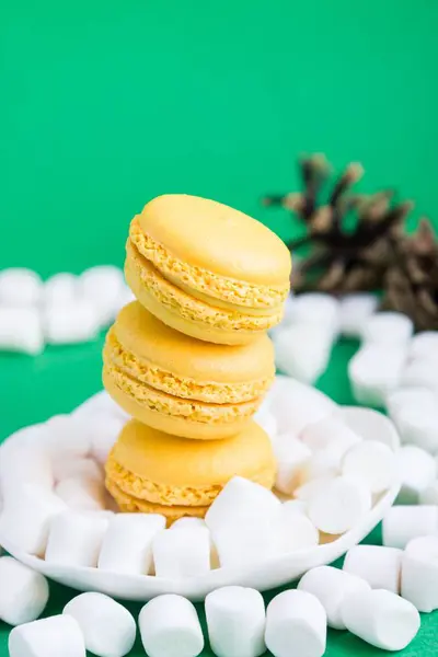 Yellow macaroons, marshmallow with pine cone on green background — Stock Photo, Image