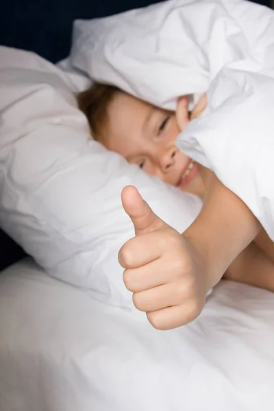 Senyum anak laki-laki terletak di tempat tidur dengan linen tempat tidur putih dan menunjukkan jempol up — Stok Foto