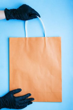 Elleri siyah ameliyat eldiveni içinde, kağıt torba tutuyor. COVID-19 'a karşı ev teslimatı önlemleri sevgiyle karşılanıyor. Hediye paketi doğrudan temas olmadan teslim ediliyor.. 